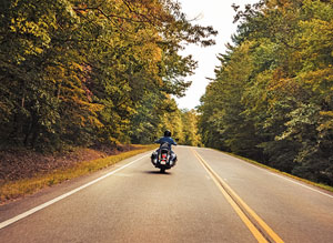 photo of the rear of a motorcyle and driver, on a road, with fall foliage.